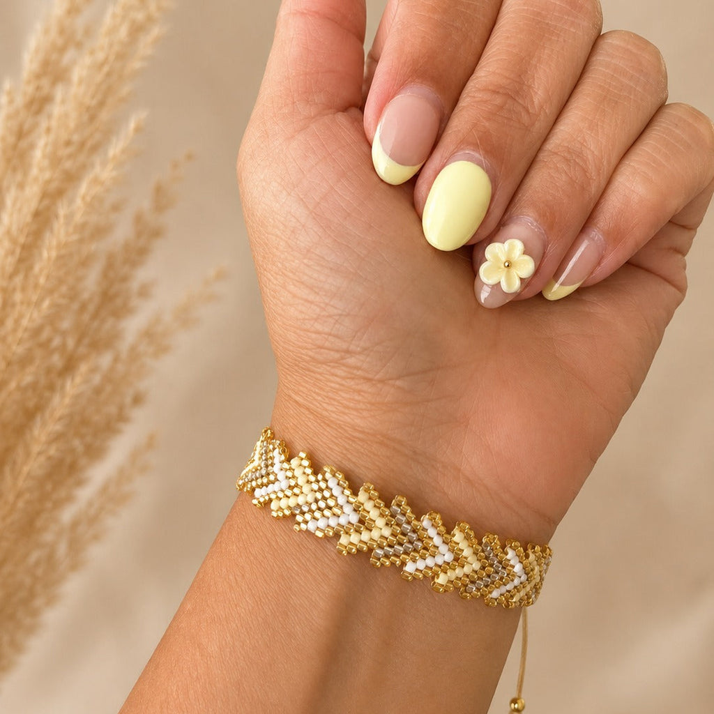 A close-up of a hand wearing a gold and white beaded chevron bracelet, with pale yellow manicured nails and a small flower accent on one nail, against a soft beige background with dried grass.