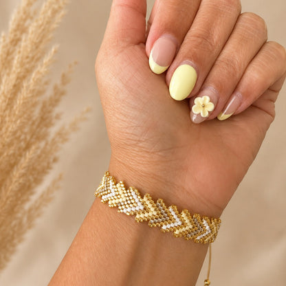 A close-up of a hand wearing a gold and white beaded chevron bracelet, with pale yellow manicured nails and a small flower accent on one nail, against a soft beige background with dried grass.