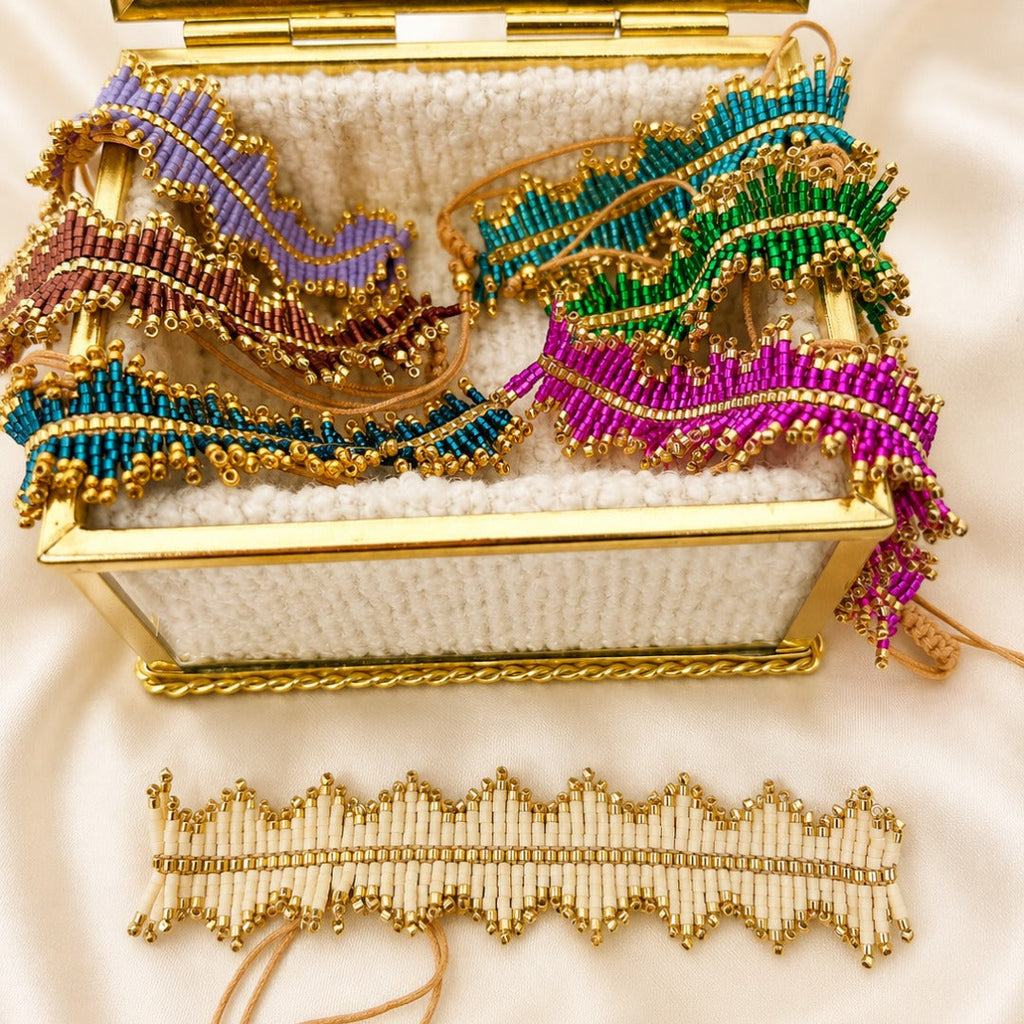 A gold-framed jewelry box lined with white fabric, filled with colorful beaded hair wraps or headbands, with a matching beaded hair accessory laid out below on a white satin background.