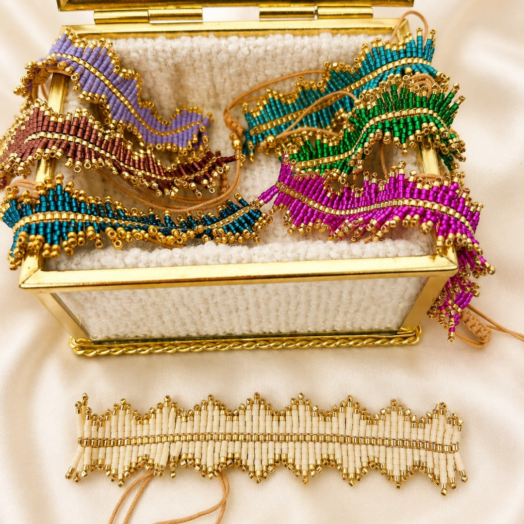 A gold-framed jewelry box lined with white fabric, filled with colorful beaded hair wraps or headbands, with a matching beaded hair accessory laid out below on a white satin background.