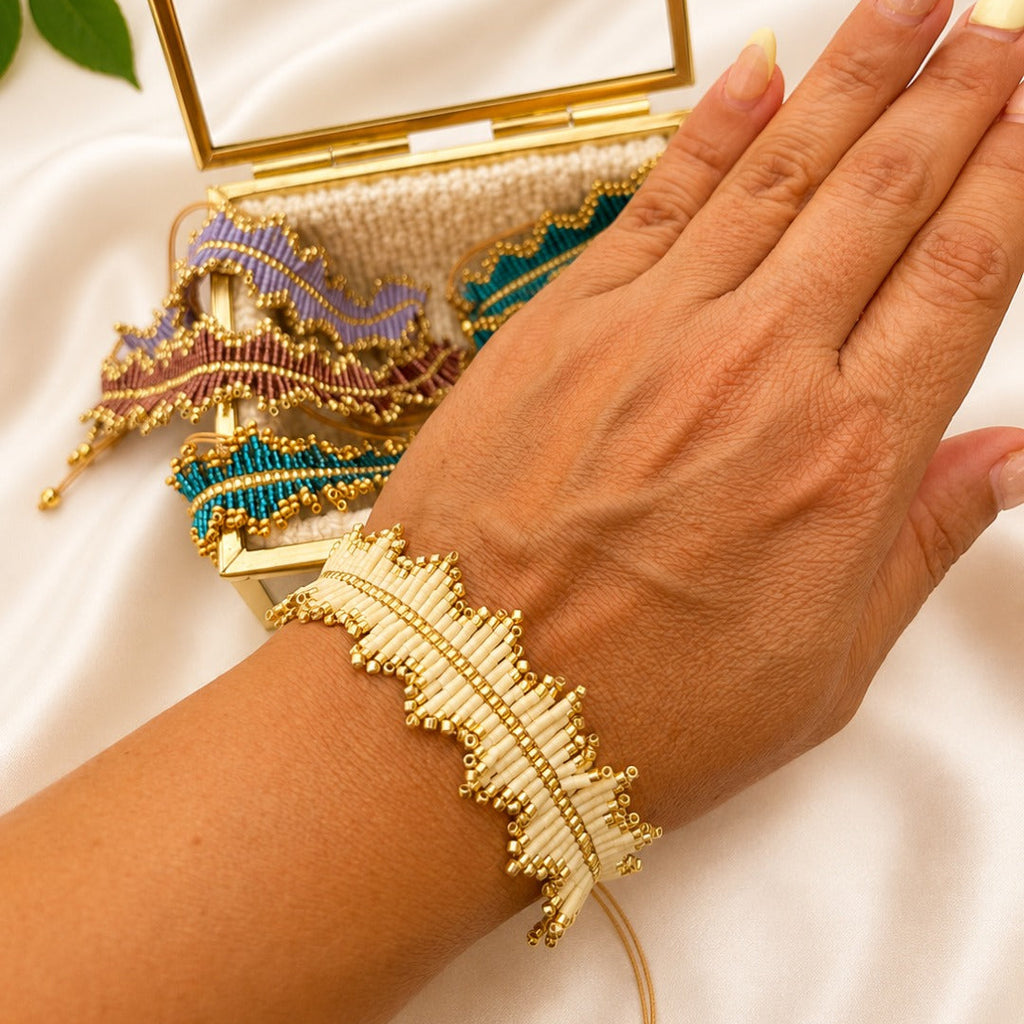 A close-up of a person's hand and wrist wearing a delicate gold and white beaded cuff bracelet, with a matching open beaded clutch bag and a gold mirror in the background on a white satin surface.