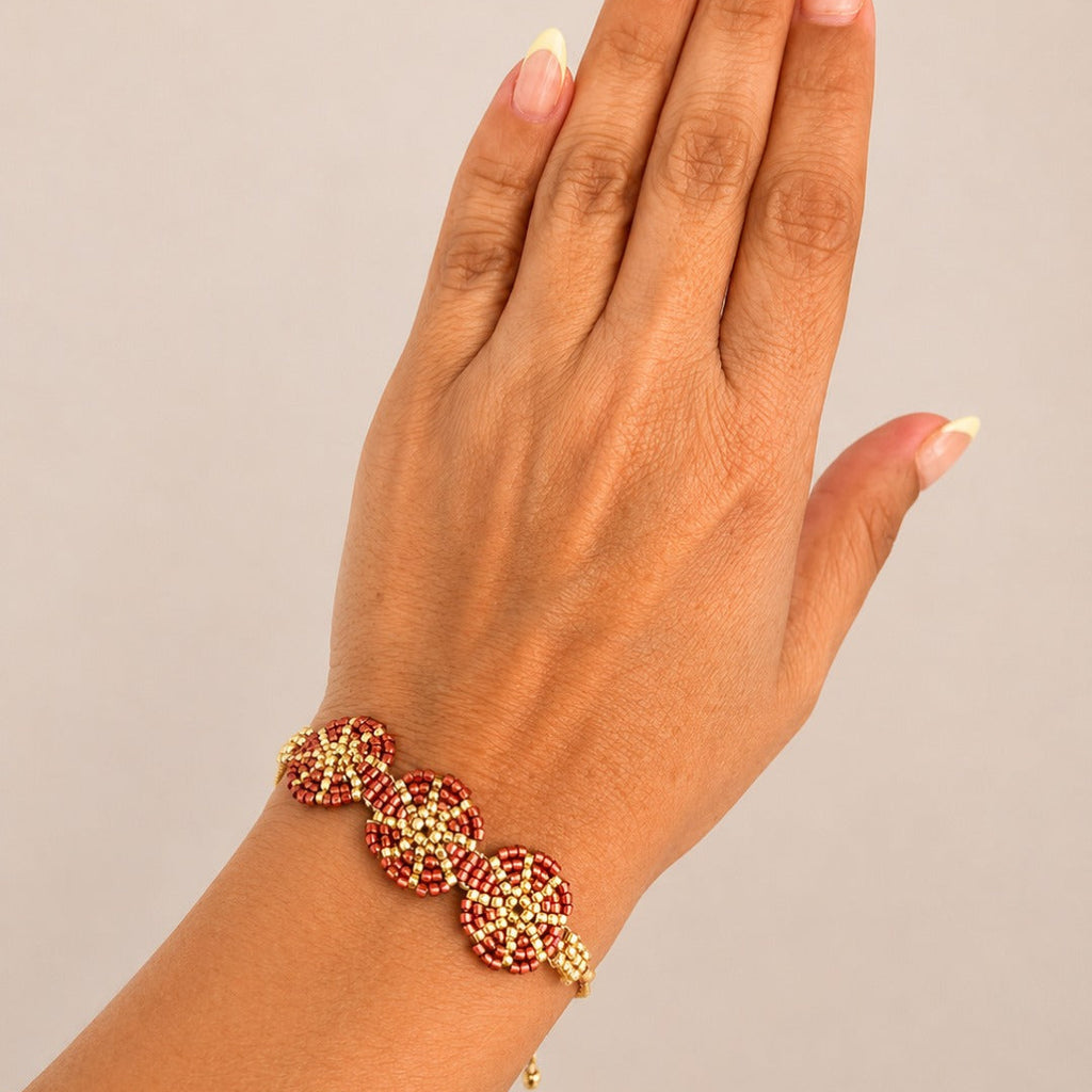 A close-up of a hand wearing a gold-plated bracelet adorned with red and gold floral beads, set against a solid light gray background.