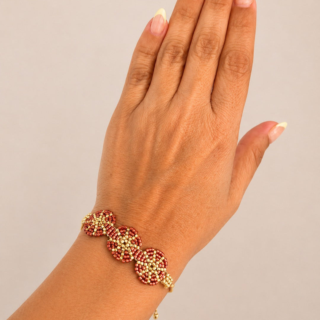 A close-up of a hand wearing a gold-plated bracelet adorned with red and gold floral beads, set against a solid light gray background.
