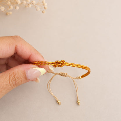 A hand holding a gold-toned beaded bracelet with an adjustable cord and knot closure, set against a soft beige background with dried flowers in the corner.