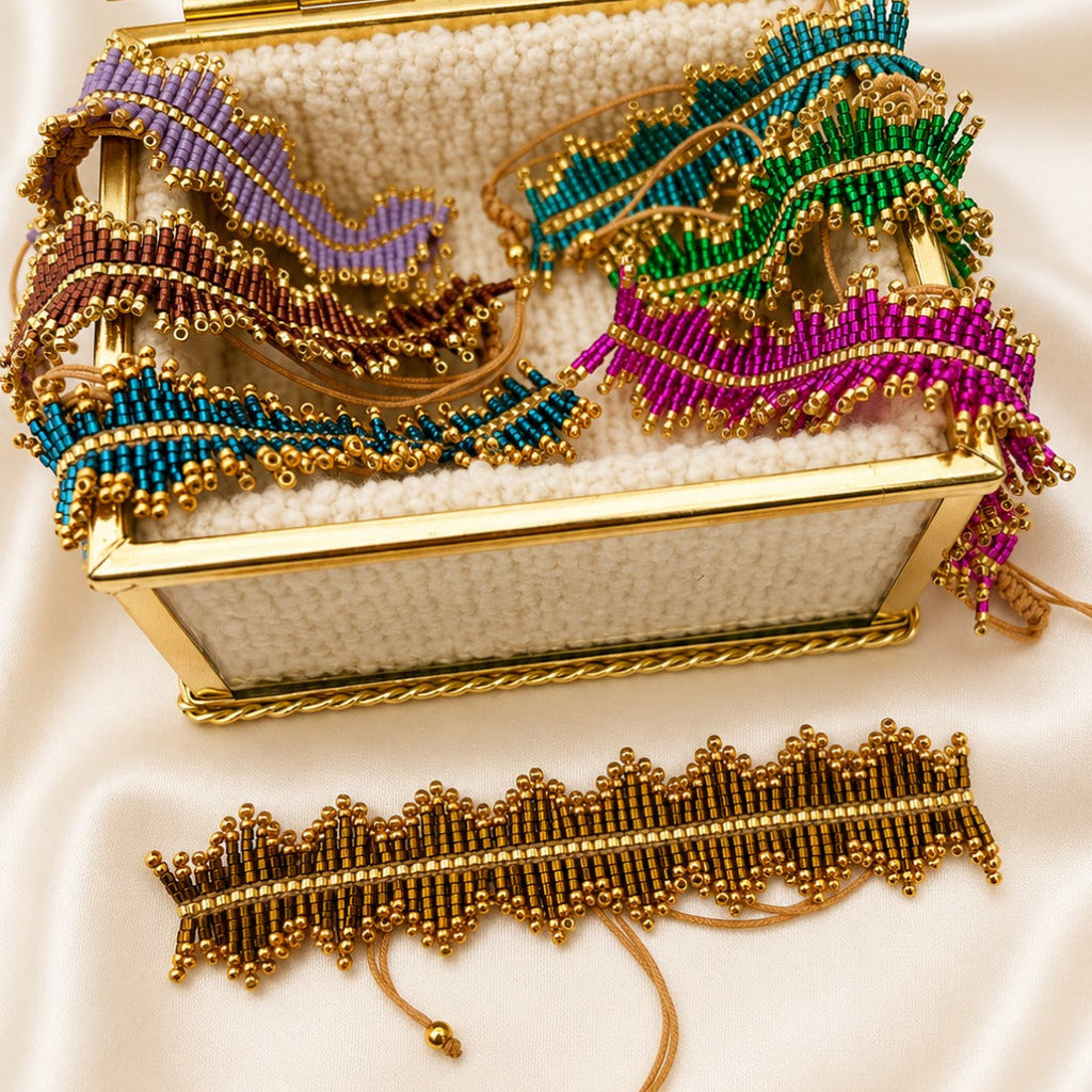 A gold-tone beaded jewelry organizer box with a clear glass lid, filled with matching beaded jewelry pieces in purple, teal, green, and magenta, displayed on a cream satin background with a matching beaded bracelet in front.