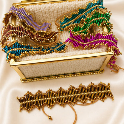 A gold-tone beaded jewelry organizer box with a clear glass lid, filled with matching beaded jewelry pieces in purple, teal, green, and magenta, displayed on a cream satin background with a matching beaded bracelet in front.