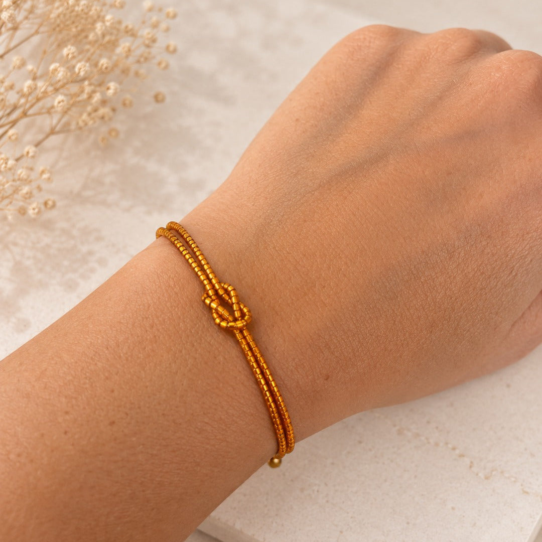 A close-up of a person's wrist wearing a thin gold-toned beaded bracelet with a knotted center detail, resting on a light textured surface with dried flowers in the background.
