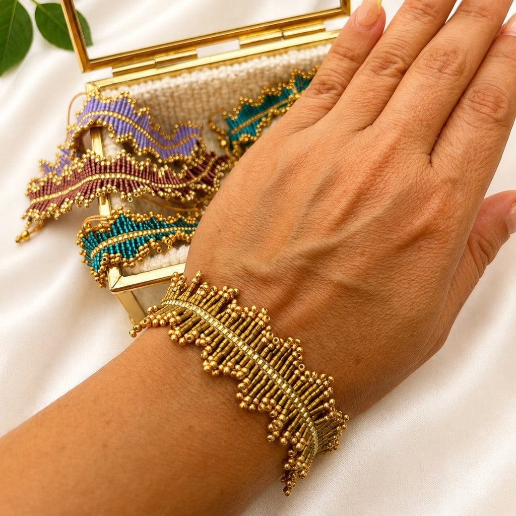 A close-up of a person's hand wearing a gold beaded wrap bracelet, with a matching gold clutch case and decorative beaded accessories in the background on a white satin surface.