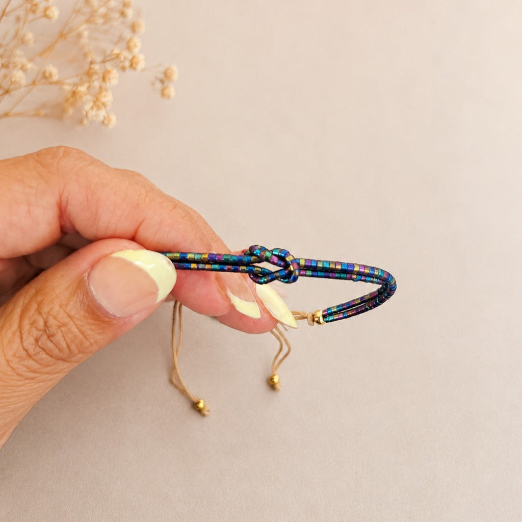A hand holding a colorful braided cord bracelet with a knot closure and gold-toned sliding adjusters, against a soft beige background with dried flowers in the corner.