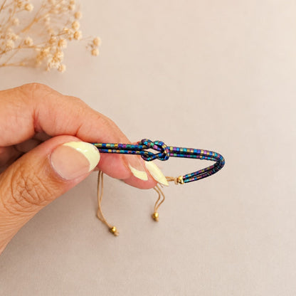 A hand holding a colorful braided cord bracelet with a knot closure and gold-toned sliding adjusters, against a soft beige background with dried flowers in the corner.