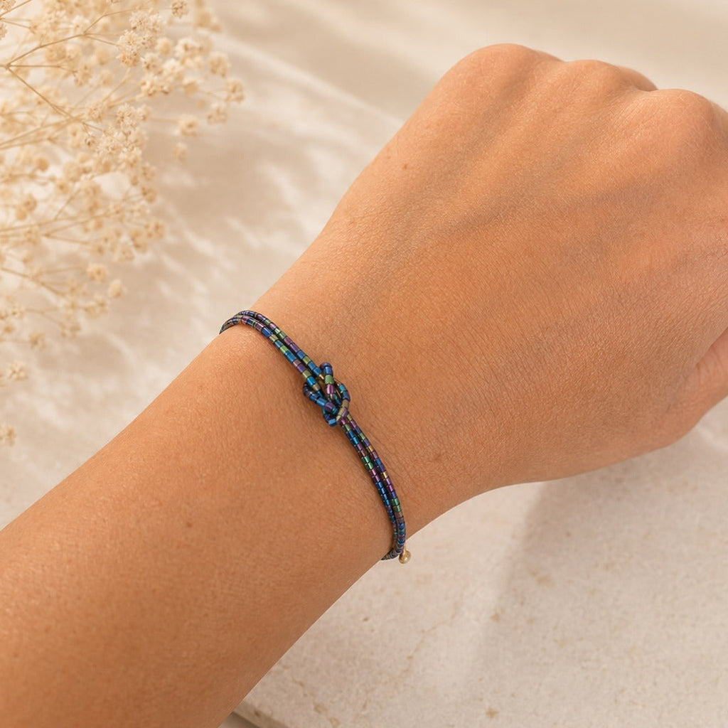 A close-up of a person's wrist wearing a delicate braided bracelet with a central knot detail. The bracelet features dark blue and red braided cords with small gold-toned accents.
