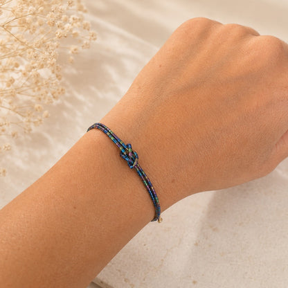 A close-up of a person's wrist wearing a delicate braided bracelet with a central knot detail. The bracelet features dark blue and red braided cords with small gold-toned accents.