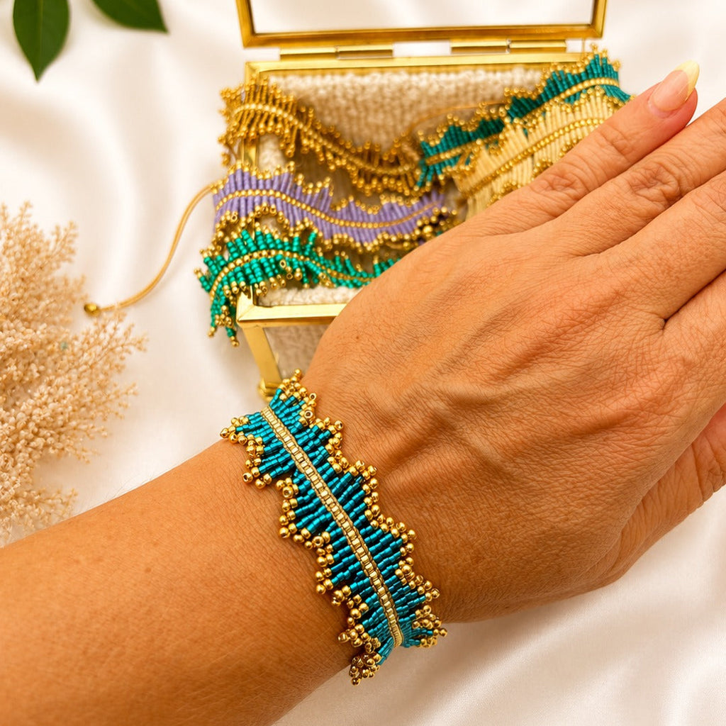 A close-up of a person's hand wearing a turquoise and gold beaded bracelet, with a matching open gold clutch bag in the background on a white satin surface.