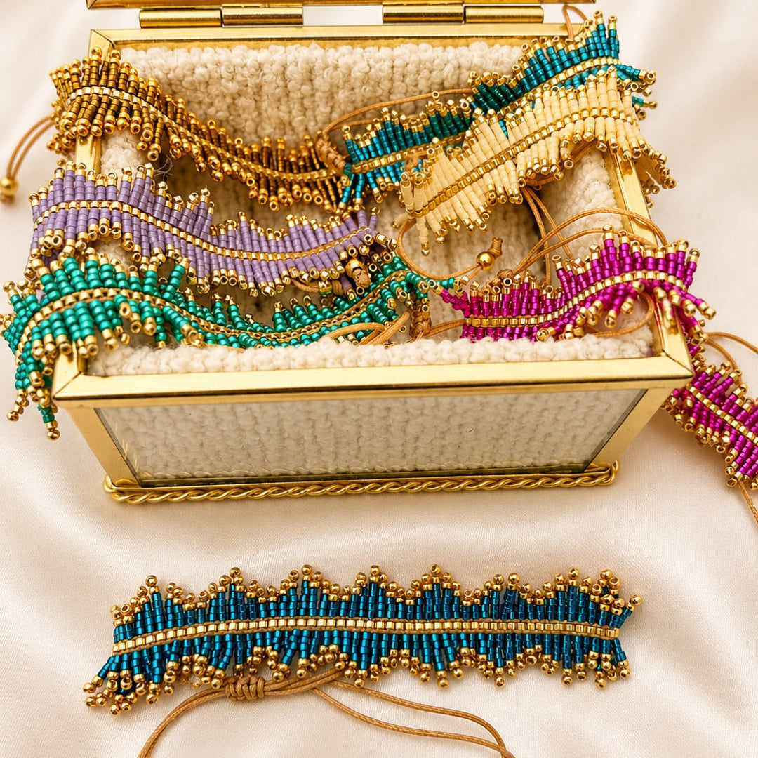 A gold-framed open jewelry box filled with colorful beaded bracelets and a matching bracelet placed in front, set on a cream satin background.