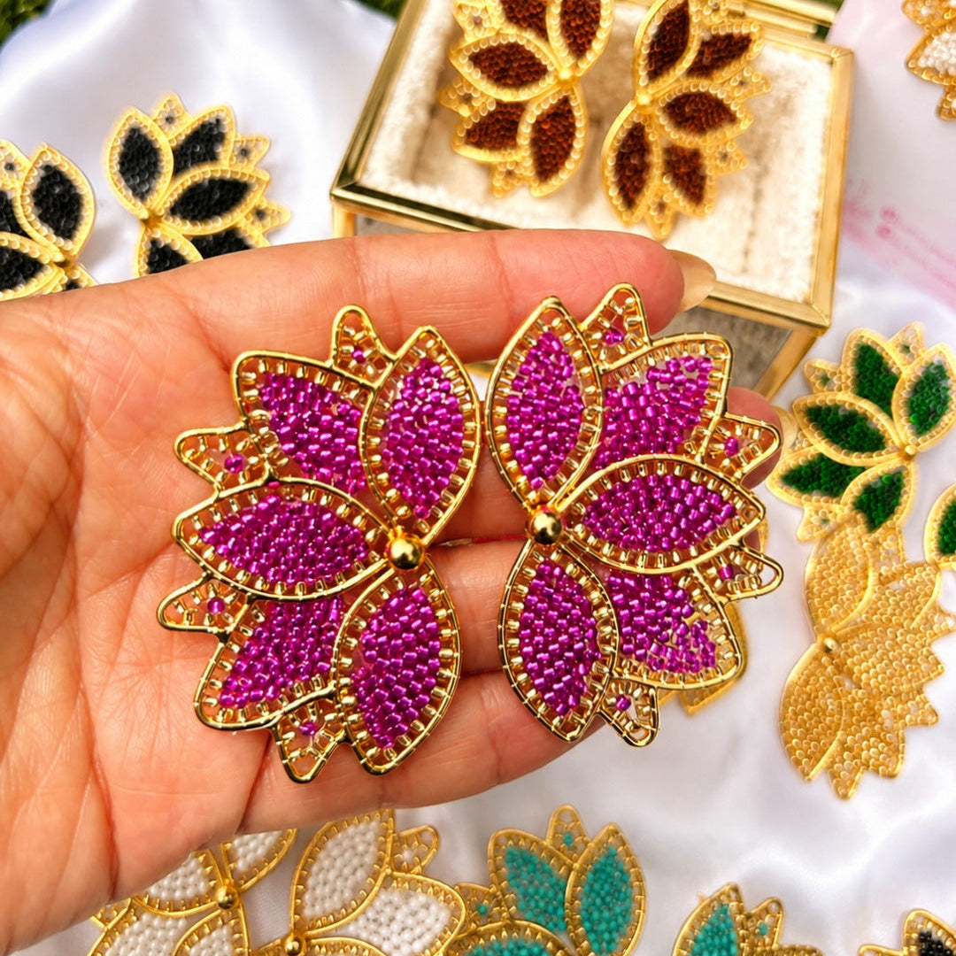 A hand holds a pair of gold-tone butterfly-shaped earrings embellished with bright pink rhinestones. Other butterfly earrings in different colors and a small gold jewelry box are arranged around them on white fabric with green grass visible in the background.