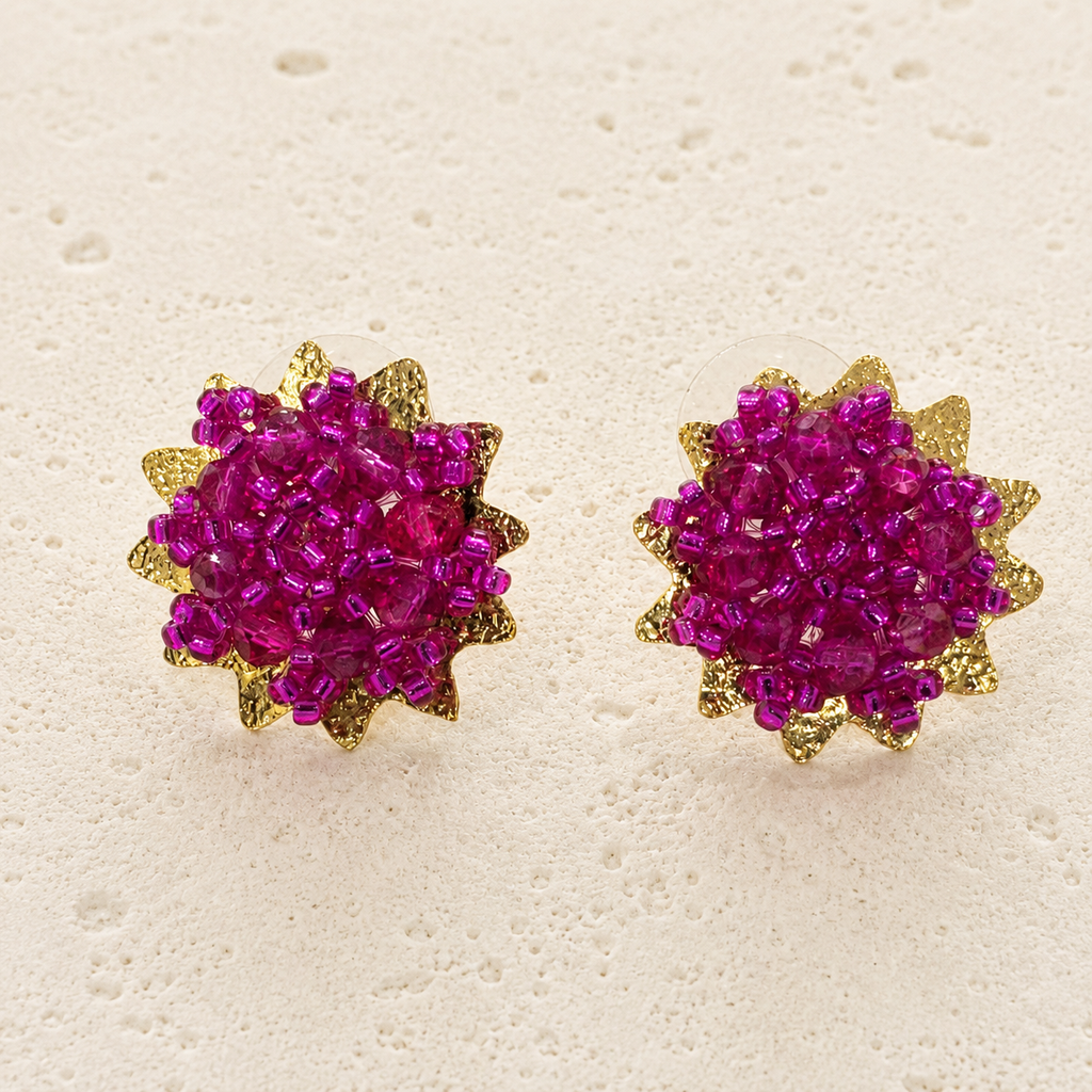 A pair of gold starburst stud earrings with vibrant magenta-pink crystal clusters in the center, displayed on a textured off-white surface.
