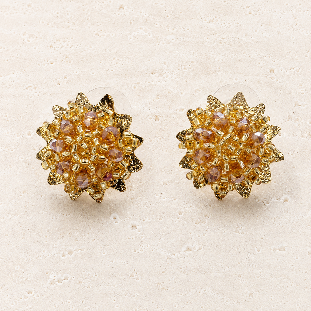 A pair of gold-toned starburst stud earrings with iridescent pinkish-purple rhinestones, displayed on a textured beige surface.