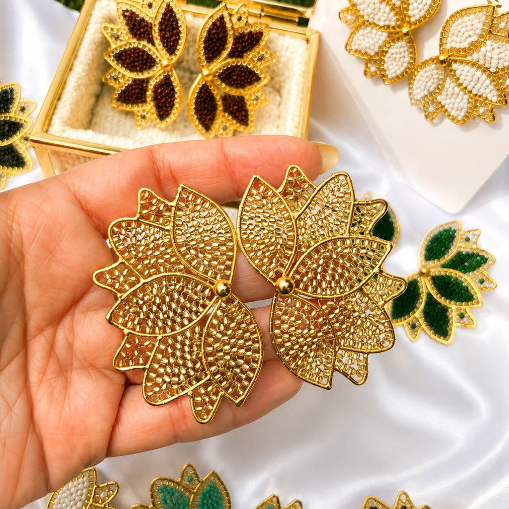 Gold-tone lotus-shaped earrings with textured rhinestone detailing, held in a hand. Other color variations of the earrings, including green, white, and black, are displayed on a white satin fabric alongside an open gold jewelry box and a branded card reading BellaJu.
