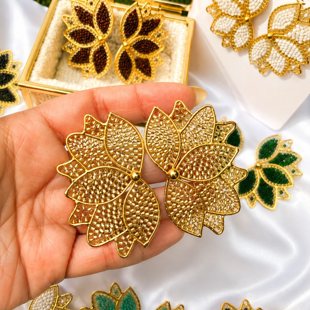 Gold-tone lotus-shaped earrings with textured rhinestone detailing, held in a hand. Other color variations of the earrings, including green, white, and black, are displayed on a white satin fabric alongside an open gold jewelry box and a branded card reading BellaJu.