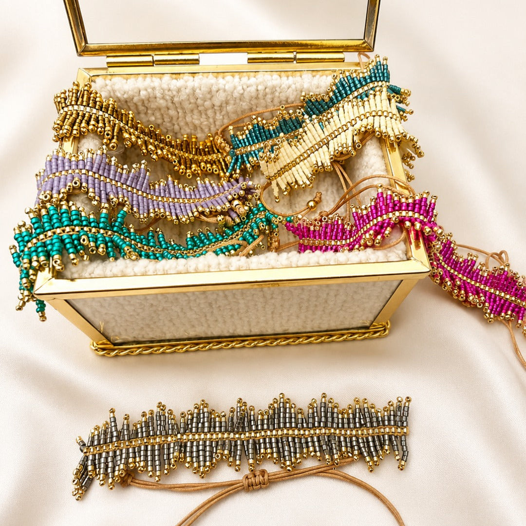 A gold-framed jewelry box filled with beaded feather-shaped accessories in teal, lavender, cream, and magenta, with one matching accessory laid out in front on a cream satin background.