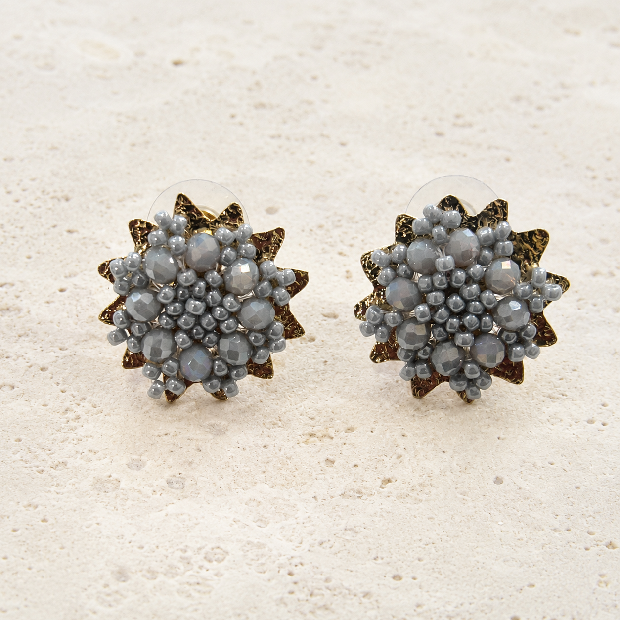 A pair of starburst stud earrings with faceted gray-blue crystals and textured bronze star-shaped metal settings, displayed on a light beige textured surface.