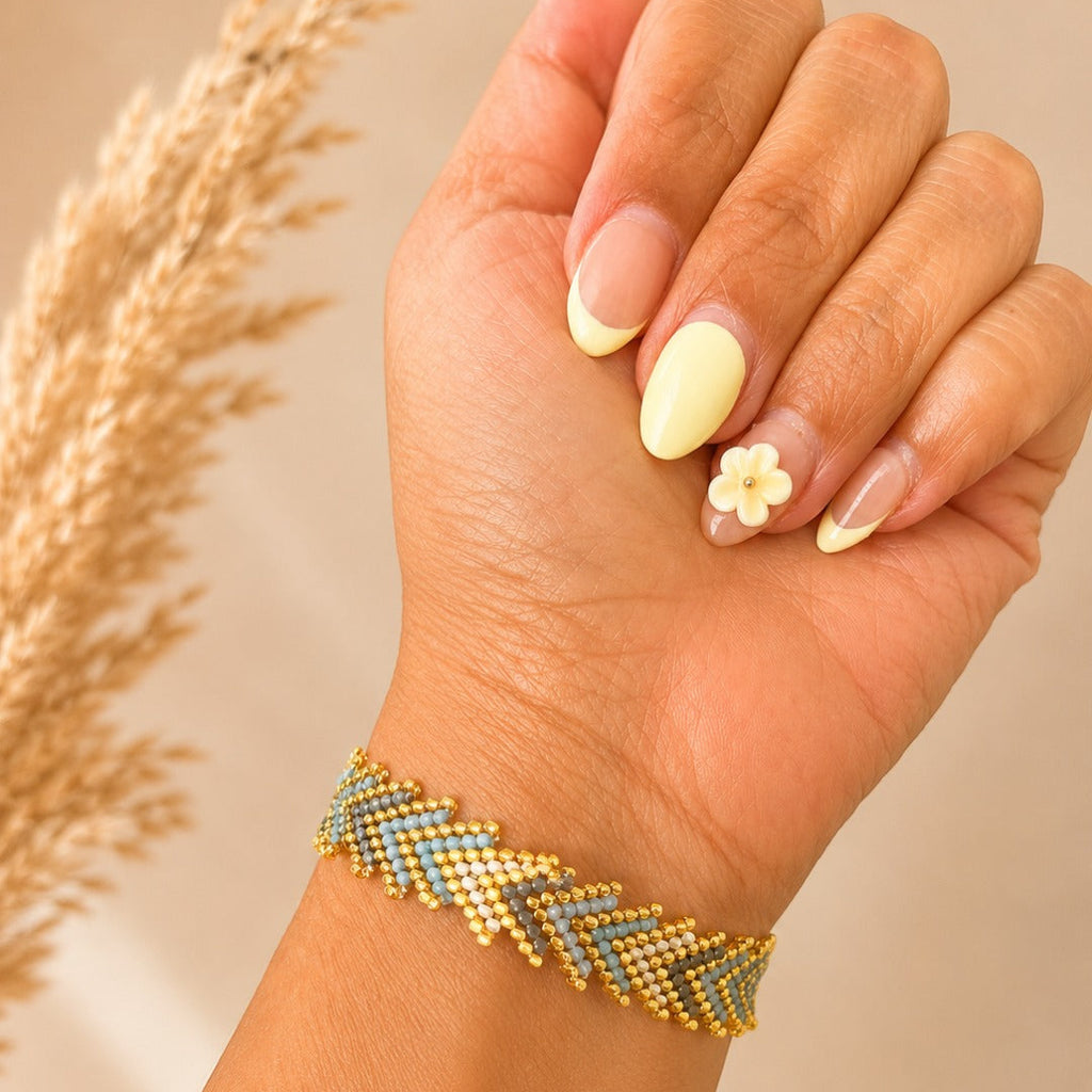 A close-up of a hand wearing a beaded chevron bracelet with gold, light blue, and gray beads, styled with a cream flower nail art accent against a warm beige background with dried grass.