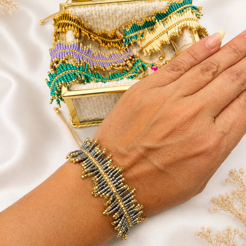 A close-up of a hand wearing a beaded leaf-shaped bracelet, with a matching open jewelry box behind it on a white satin background.