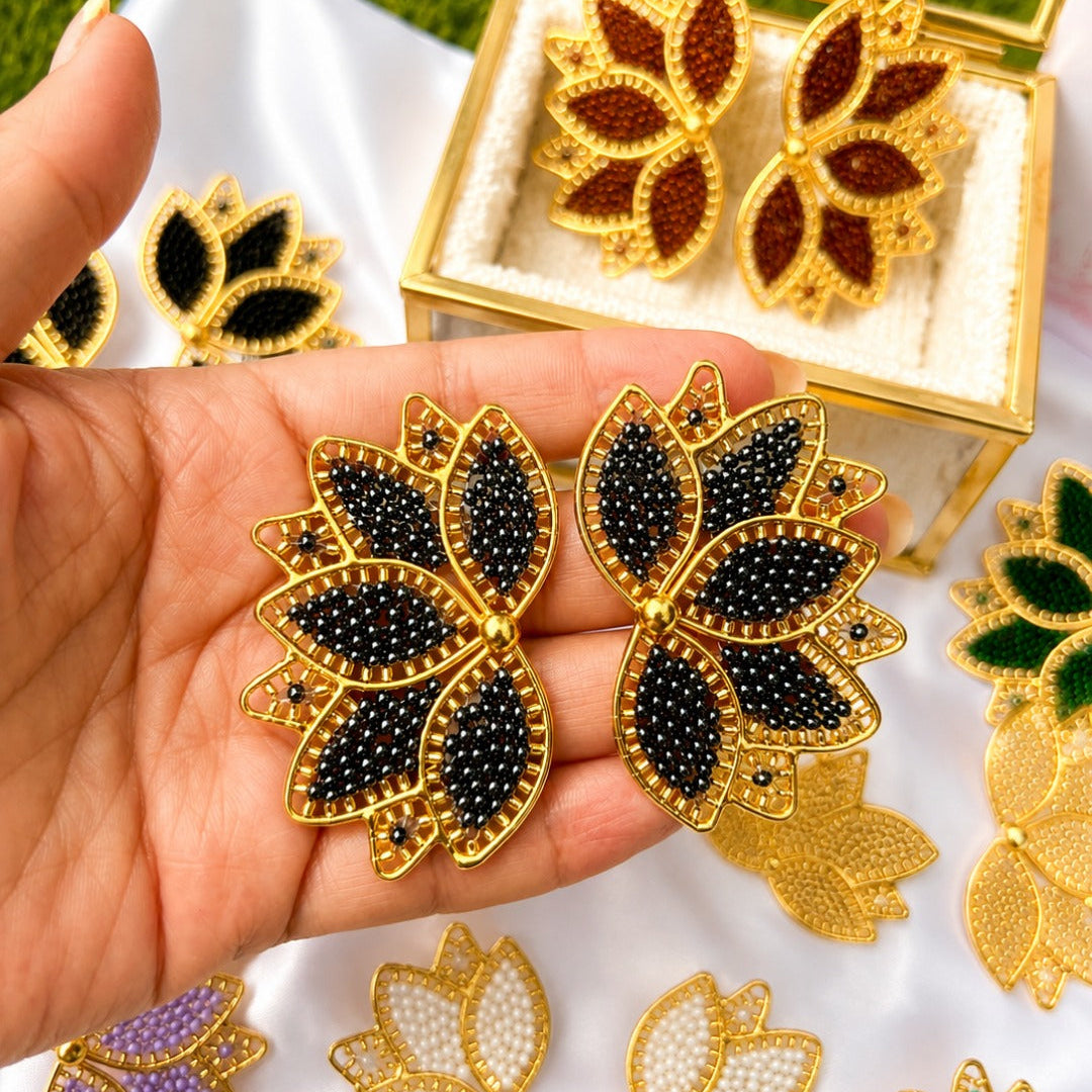 Gold-tone lotus-shaped earrings with black and white beadwork, held in a hand over a white fabric surface with other color variants and an open display box in the background.