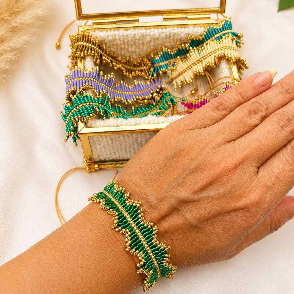 A close-up of a person's hand wearing a green and gold beaded leaf-shaped bracelet, with a matching open gold clutch case behind it on a white fabric background.