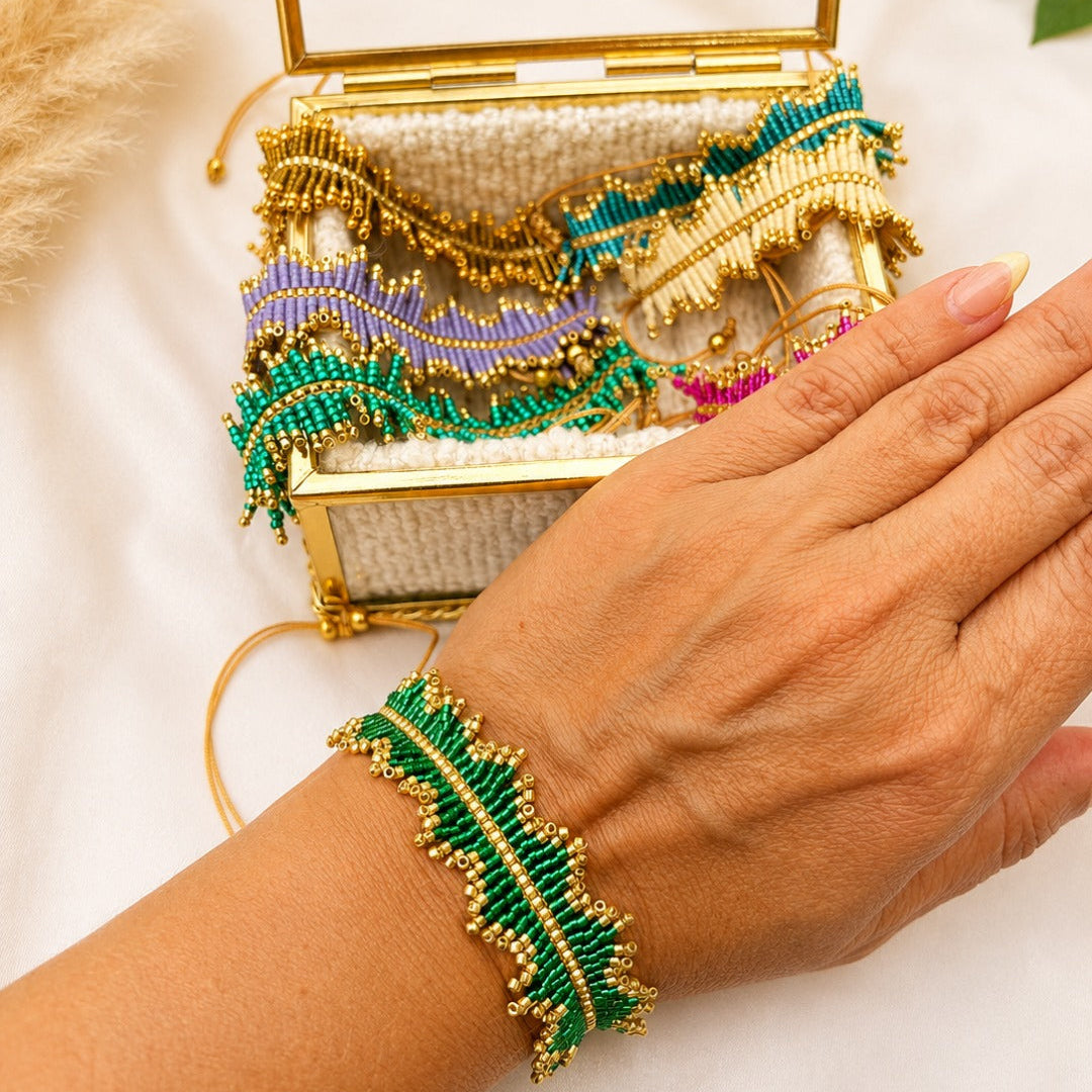 A close-up of a person's hand wearing a green and gold beaded leaf-shaped bracelet, with a matching open gold clutch case behind it on a white fabric background.