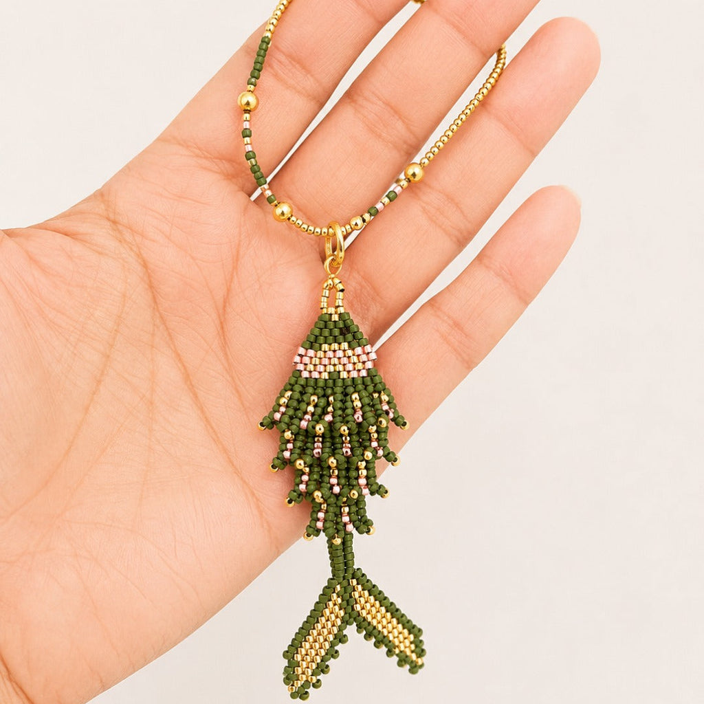 A handmade beaded fish pendant necklace held in a hand. The necklace features a gold-tone chain and a fish-shaped pendant made of green, gold, and light pink seed beads.