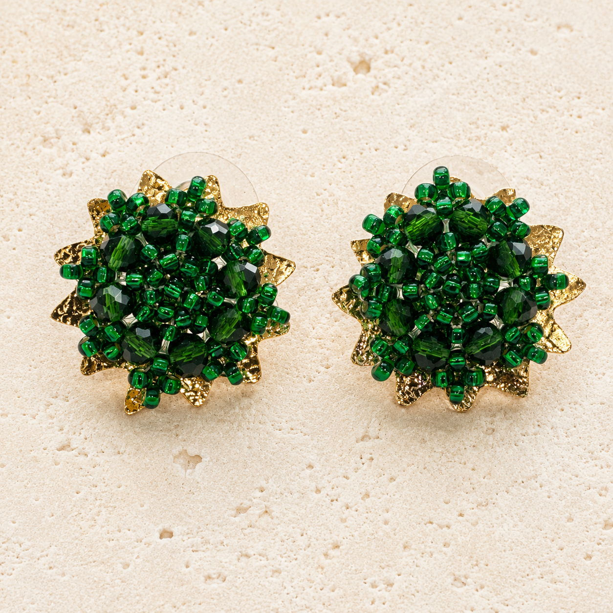 A pair of round green beaded flower stud earrings with gold-toned accents, displayed on a textured beige surface.
