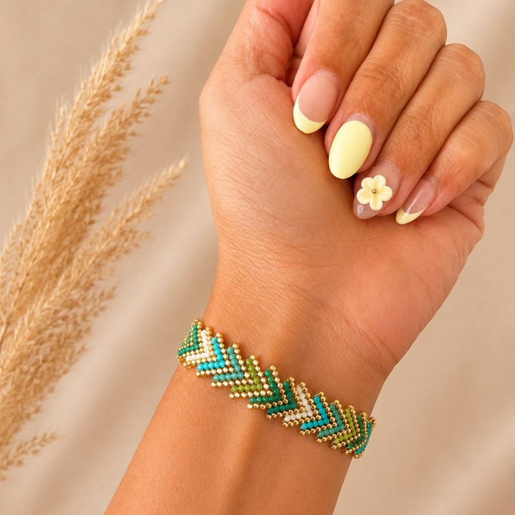 A close-up of a person's hand and wrist wearing a beaded chevron bracelet in green, teal, and gold. The hand has pale yellow manicured nails with a small flower accent, and dried grass is visible on the left against a soft beige background.