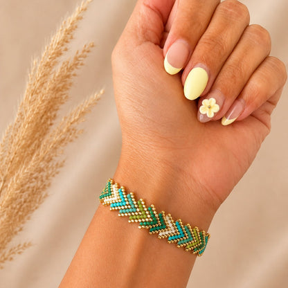 A close-up of a person's hand and wrist wearing a beaded chevron bracelet in green, teal, and gold. The hand has pale yellow manicured nails with a small flower accent, and dried grass is visible on the left against a soft beige background.