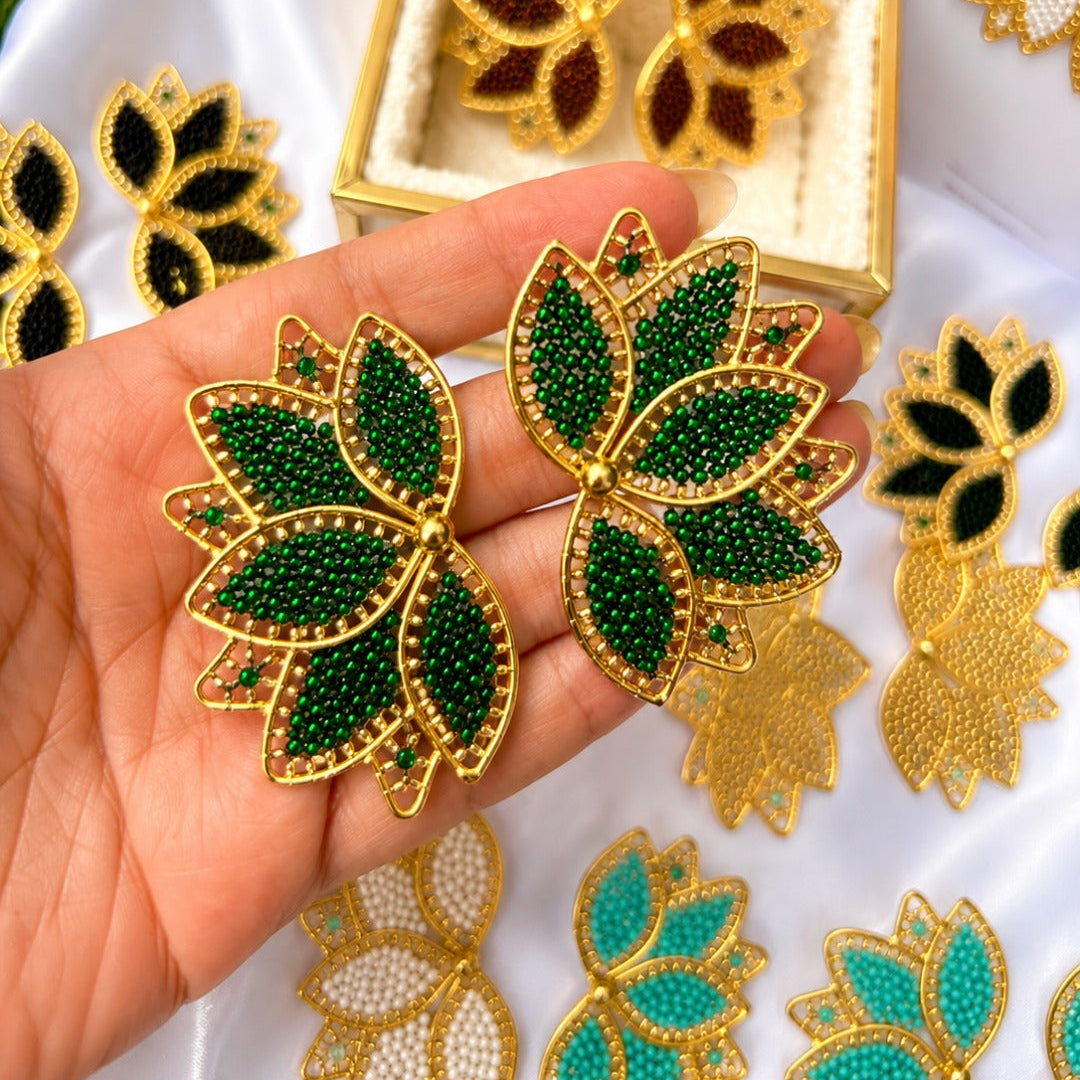 A hand holds a pair of gold-tone leaf-shaped earrings embellished with dark green seed beads, with a matching open display box and additional leaf jewelry pieces arranged on white fabric in the background.
