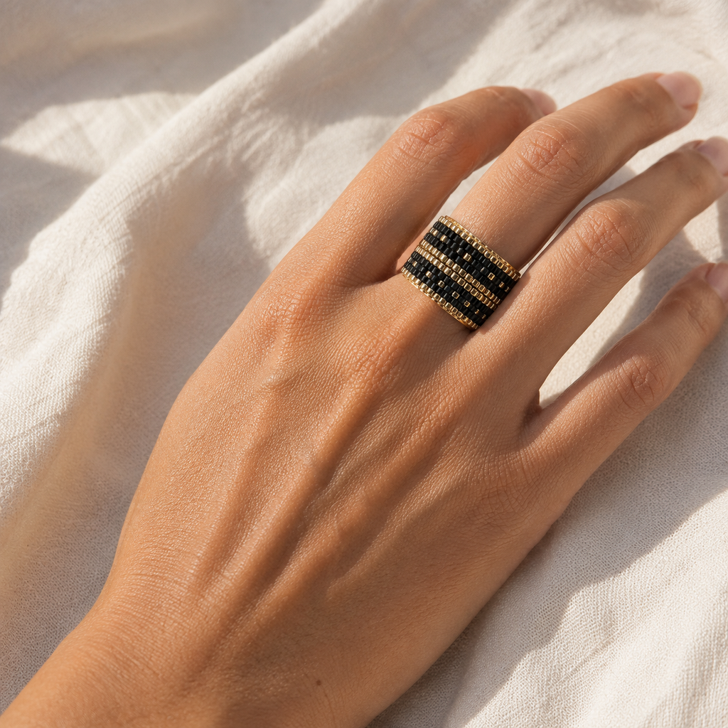 A close-up of a hand wearing a gold ring with black enamel inlay and small white stone accents, resting on a textured white fabric background.