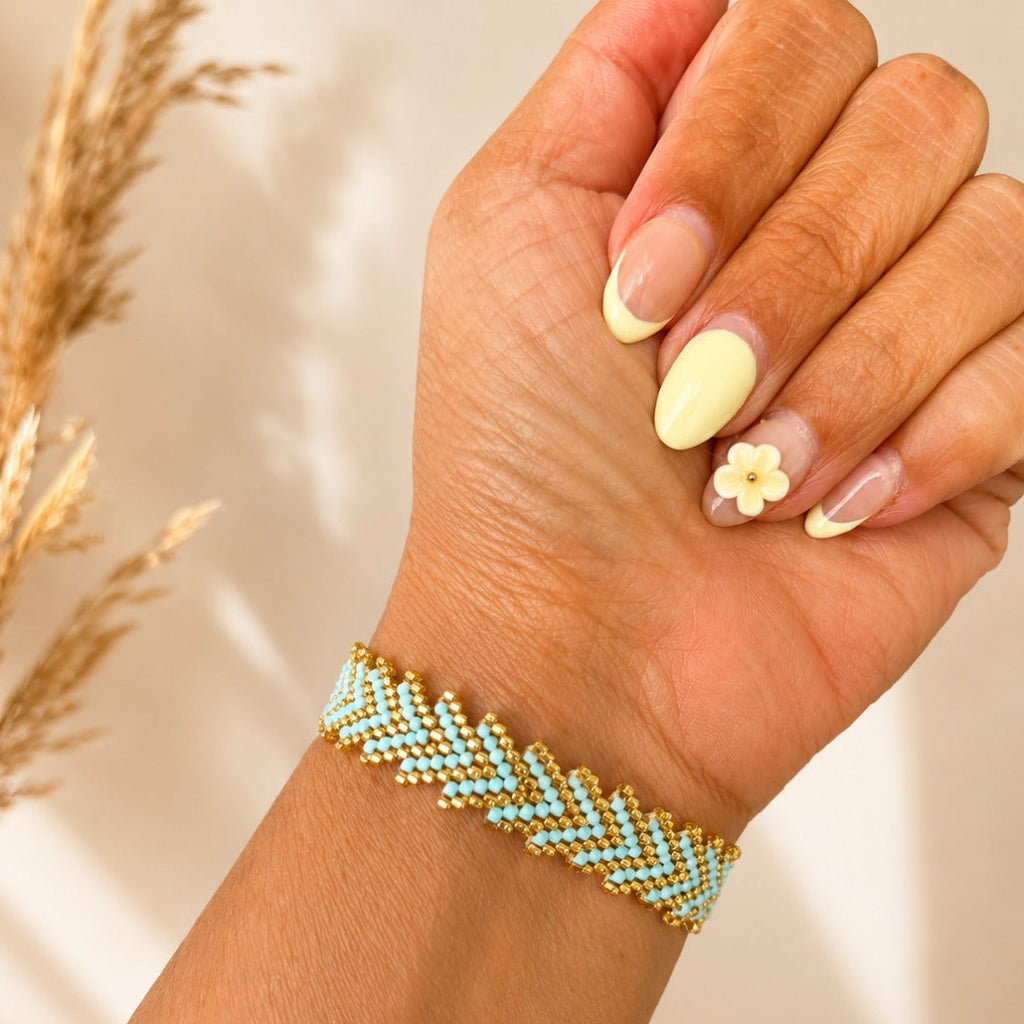 A close-up of a hand wearing a beaded chevron bracelet with light blue and white beads outlined in gold. The hand has pale yellow and white nail art, and the background is a warm beige with soft dried grass elements.