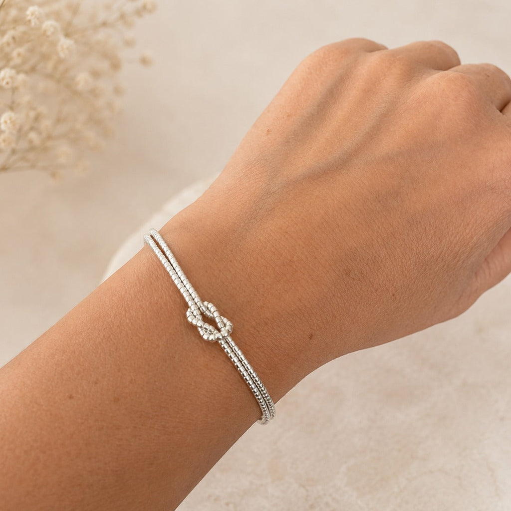 A silver-toned double-strand beaded bracelet with a central knot design, shown worn on a person's wrist against a soft beige background with dried flowers.