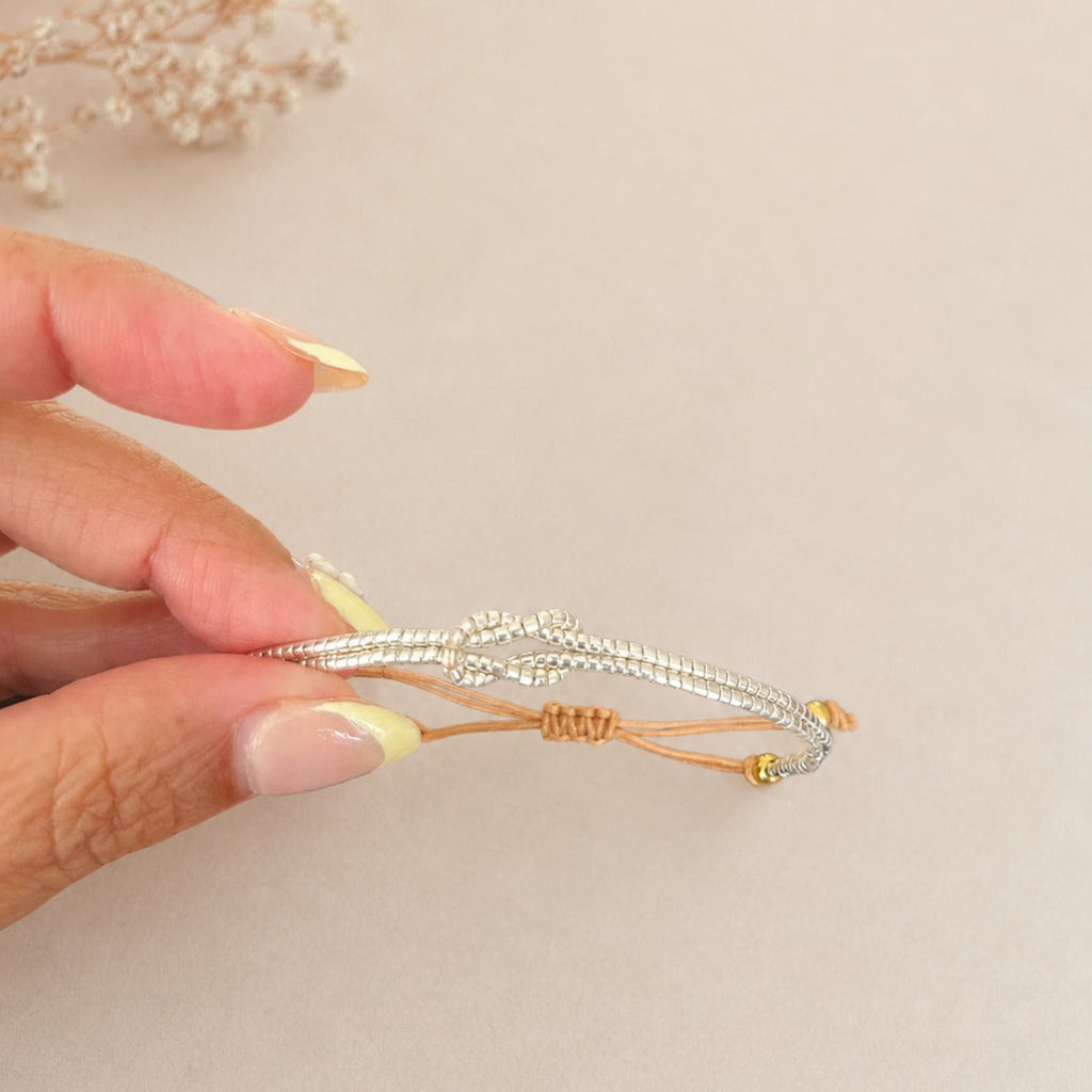 A hand with pale yellow manicured nails holds a delicate silver-toned beaded bracelet with a gold accent cord against a soft beige background with dried flowers in the corner.
