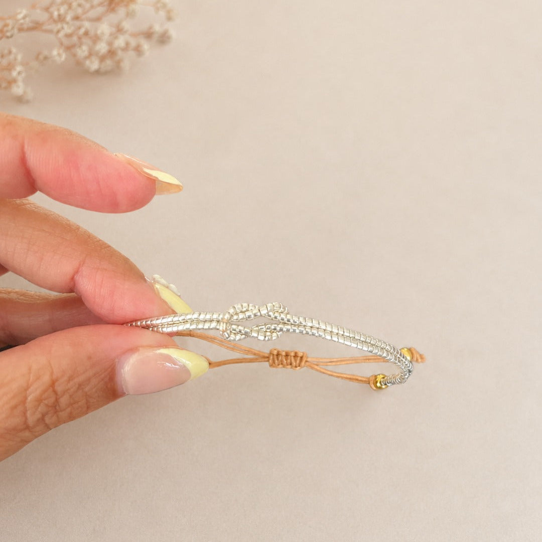 A hand with pale yellow manicured nails holds a delicate silver-toned beaded bracelet with a gold accent cord against a soft beige background with dried flowers in the corner.