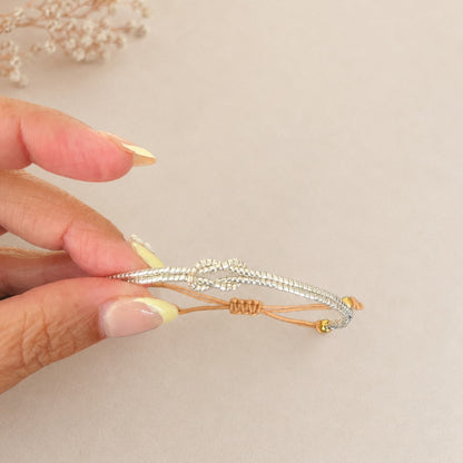 A hand with pale yellow manicured nails holds a delicate silver-toned beaded bracelet with a gold accent cord against a soft beige background with dried flowers in the corner.