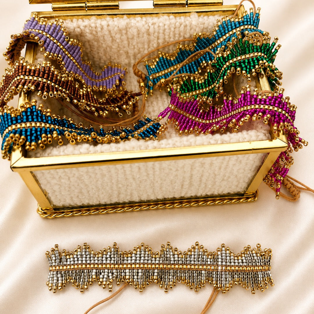 A gold-framed mirror box filled with colorful beaded accessories, including a blue and purple beaded wrap, a green and teal beaded wrap, and a magenta beaded wrap, with a matching silver and gold beaded choker displayed below on a white satin background.