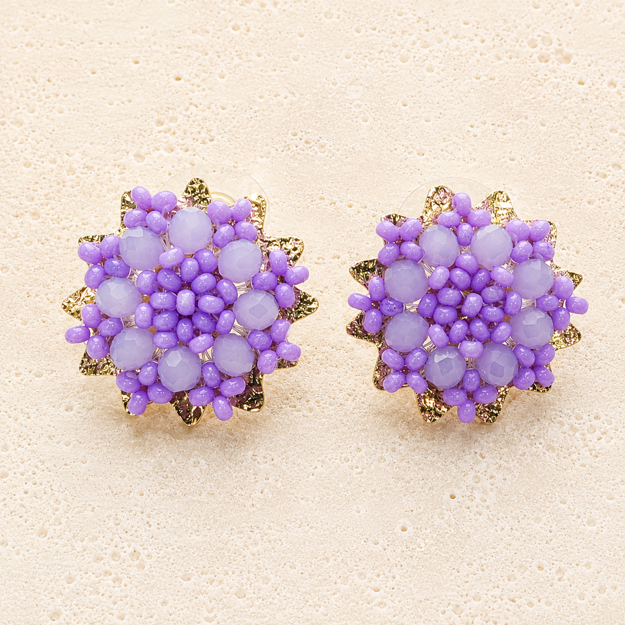 A pair of round cluster stud earrings featuring lavender purple beads and gold-tone metal accents, displayed on a textured beige surface.