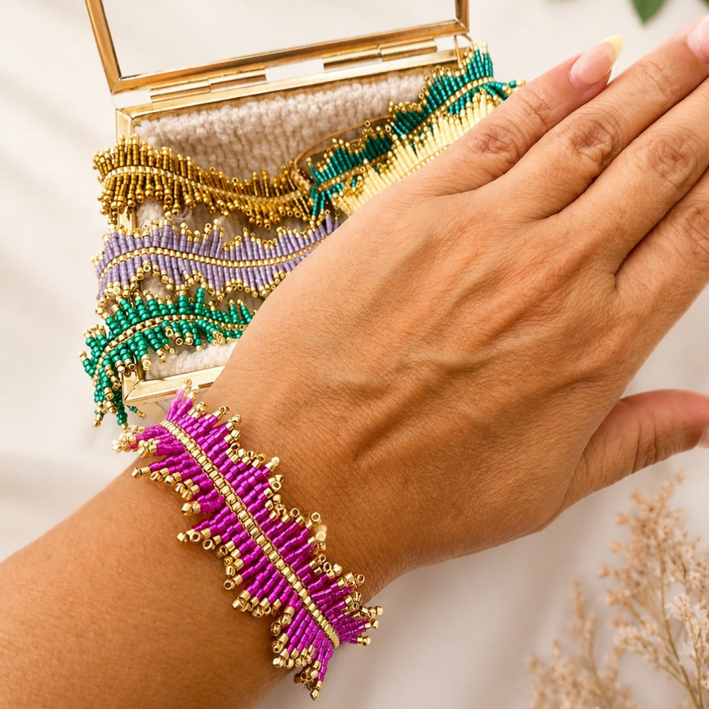 A close-up of a person's hand and wrist wearing a bright magenta and gold beaded bracelet, with a matching multicolor beaded clutch bag held behind the hand.