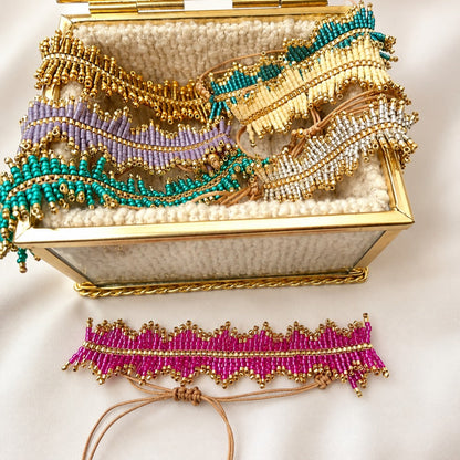 A set of handmade beaded jewelry pieces, including a gold-tone choker with purple beads, turquoise beaded wavy bands, and a pink beaded choker with tan cords, displayed in an open gold jewelry box on a white fabric background.