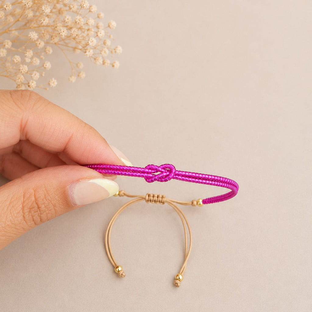 A hand with manicured nails holds a magenta pink braided cord bracelet with a knot closure and gold-plated wire loop details against a neutral beige background with dried baby's breath flowers in the corner.