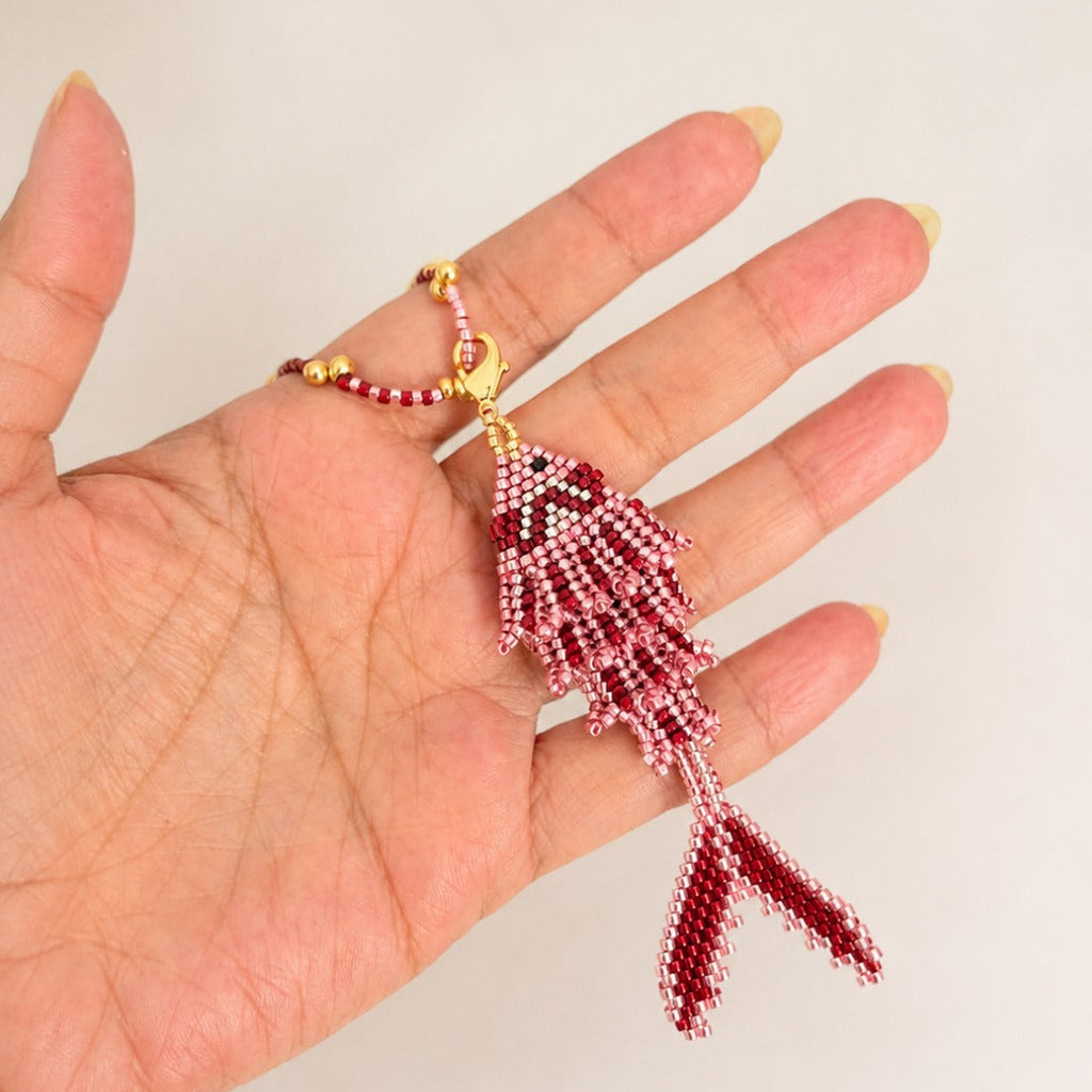 A hand holding a small beaded fish-shaped pendant or charm in red and white beads with gold-tone metal accents, against a plain light background.