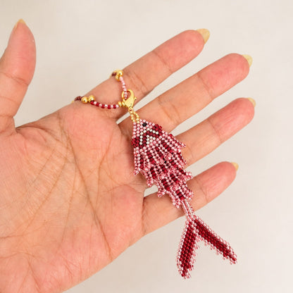 A hand holding a small beaded fish-shaped pendant or charm in red and white beads with gold-tone metal accents, against a plain light background.