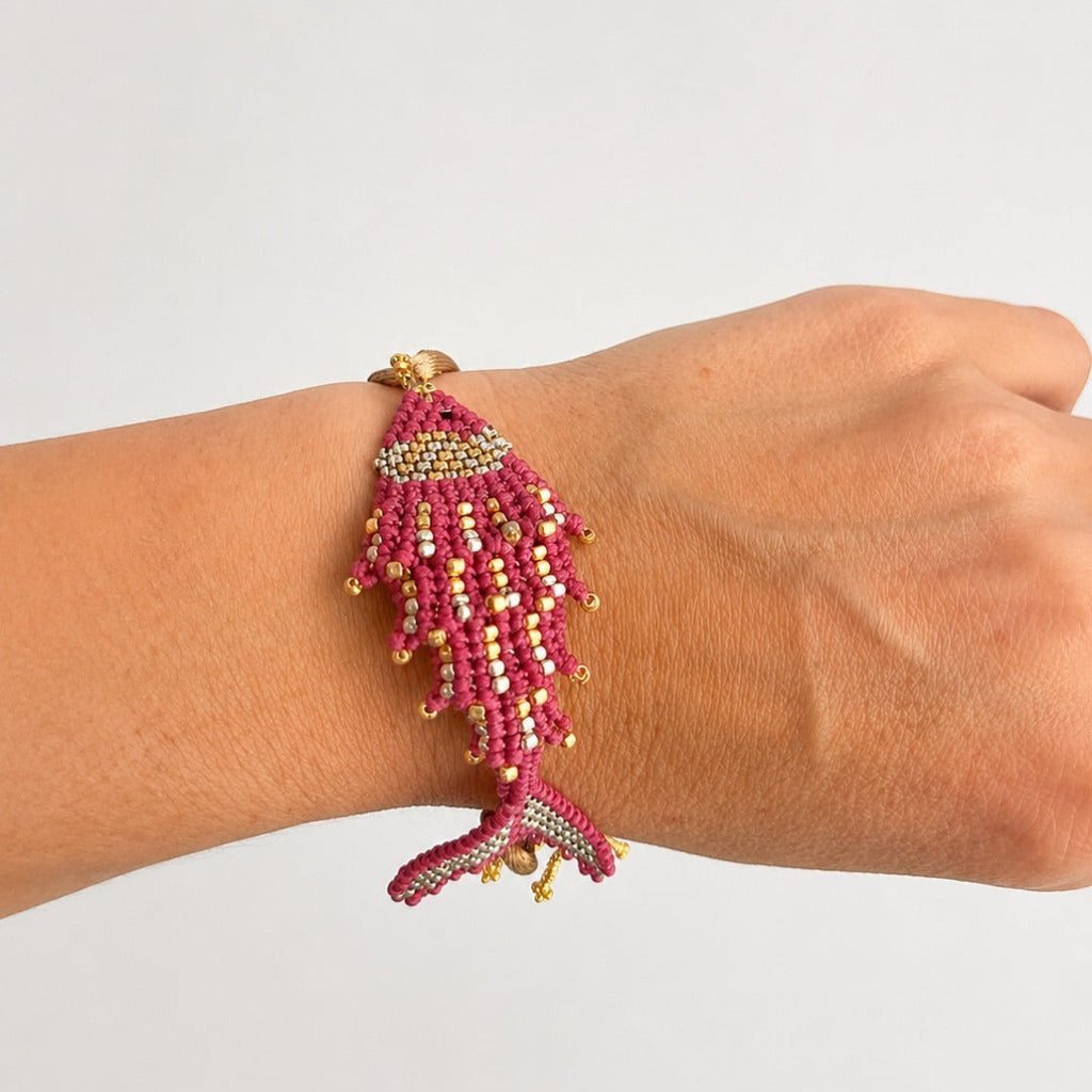 A beaded fish-shaped bracelet in pink, gold, and white, worn on a person's wrist against a plain light gray background.