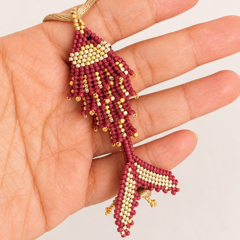 A hand holding a beaded fish-shaped ornament or charm in burgundy and cream with small gold accents, against a plain light background.